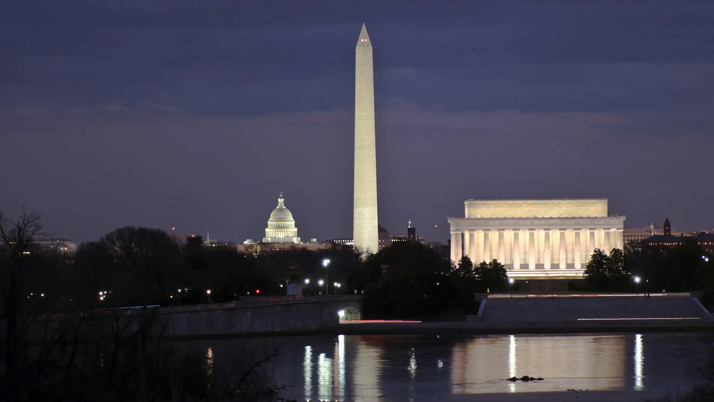 Travel-Banner Night view of Washington, D.C., featuring the illuminated Washington Monument centered, the U.S. Capitol with its dome glowing on the left, and the Lincoln Memorial brightly lit on the right.