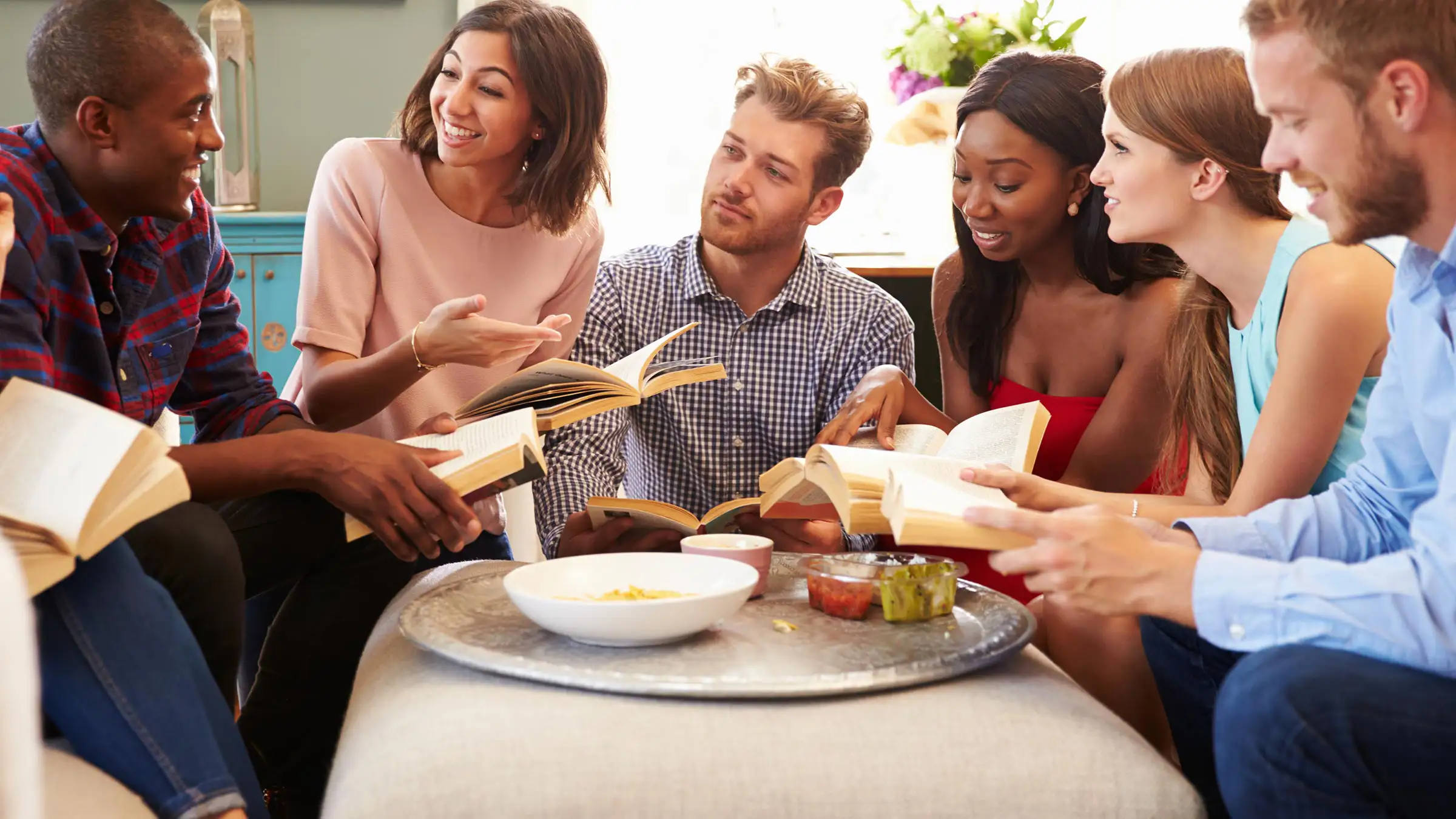Book-Club-Banner A diverse group of six people sitting in a circle in a cozy room, each holding a book. They are engaged in a lively discussion, smiling and gesturing. A large tray with snacks is on the table in the center.